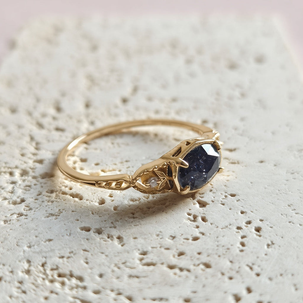 A gold ring with a dark blue faceted gemstone and a small clear accent stone, resting on a textured beige stone block against a soft pink background.